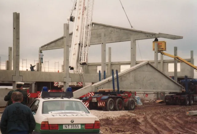 Ein Kran hebt Beton-Dachbinder auf den Rohbau, Männer schauen zu